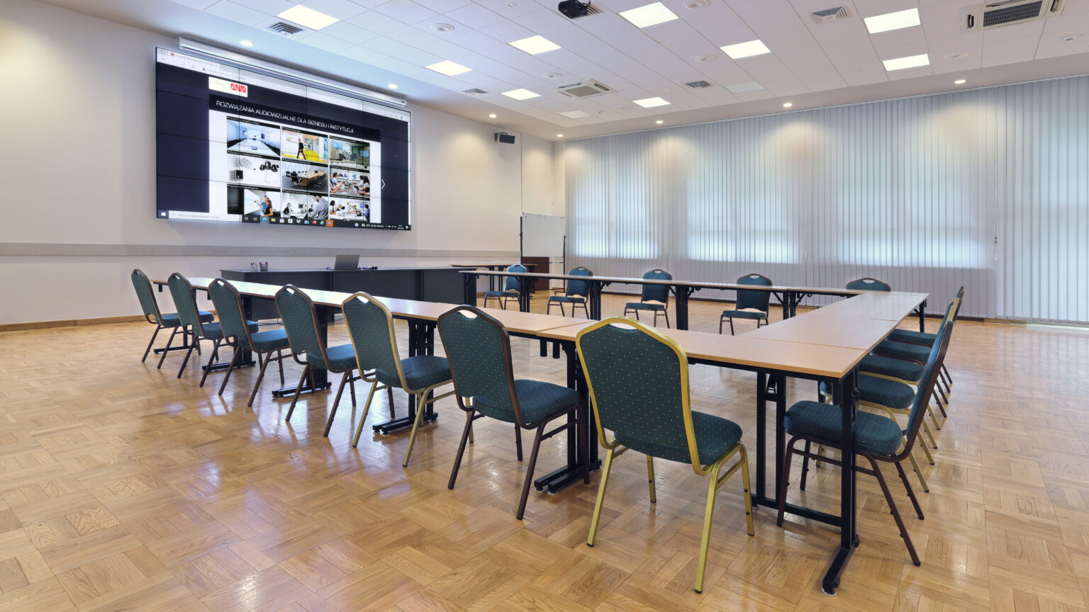 Sala wykładowa Krajowej Szkoły Skarbowości w Otwocku ze stołami w podkowę i ścianą wizyjną