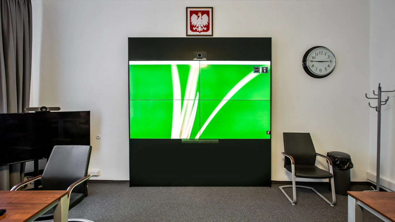 Ściana wideo 2x2 w czarnej zabudowie z kamerą nad monitorami, w sali Urzędu Marszałkowskiego Województwa Małopolskiego; nad nią godło, obok zegar, po lewej mobilny monitor i krzesła