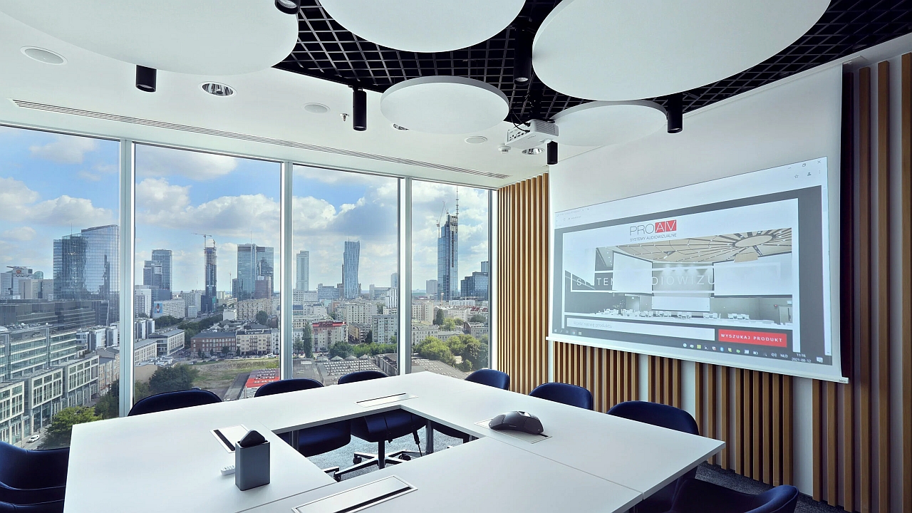Sala konferencyjna w biurze GATX Rail Poland z przeszkloną ścianą i widokiem na wieżowce, białym stołem, ekranem projekcyjnym na ścianie z drewnianymi lamelami i okrągłymi panelami akustycznymi pod sufitem