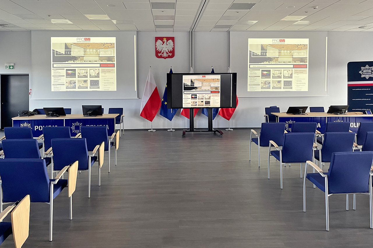 Sala odpraw z rzędami krzeseł, dwa ekrany projekcyjne na ścianie i monitor interaktywny między flagami