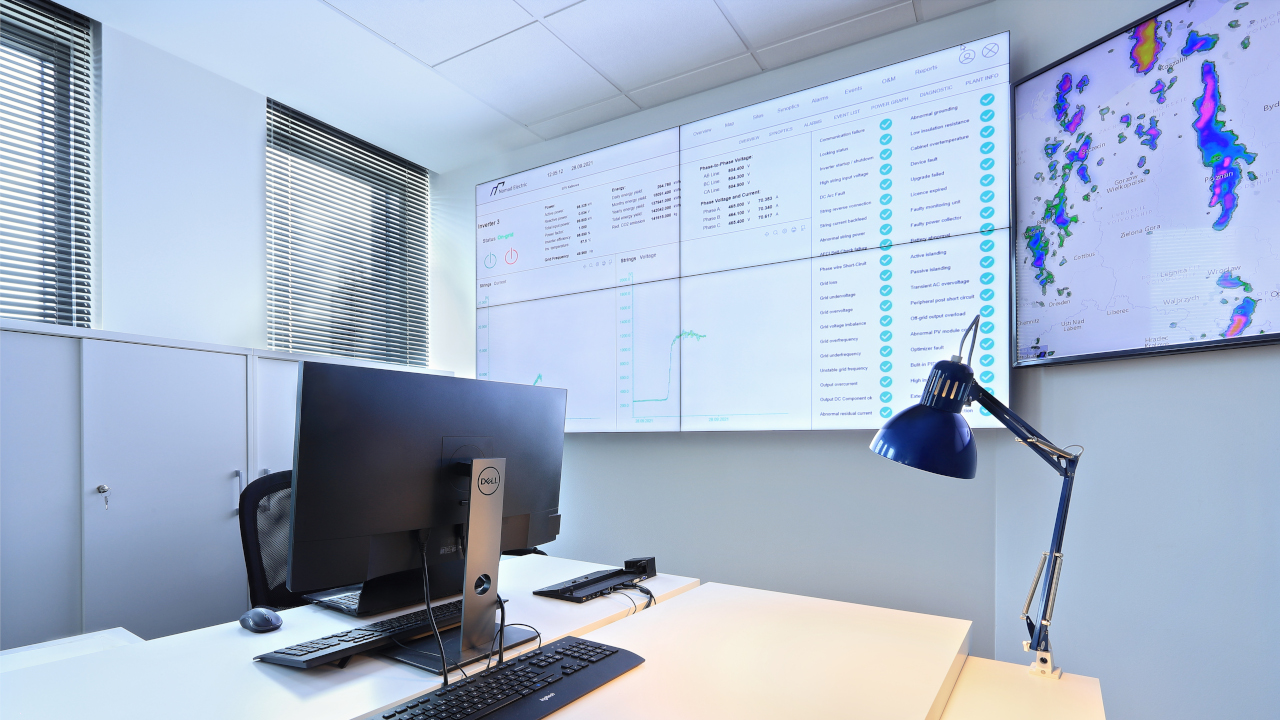 Control room Nomad Electric: ściana wideo 2x2 z panelem wskaźników instalacji fotowoltaicznej, obok monitor z mapą radarową opadów, na pierwszym planie biurko operatora z komputerem i lampką