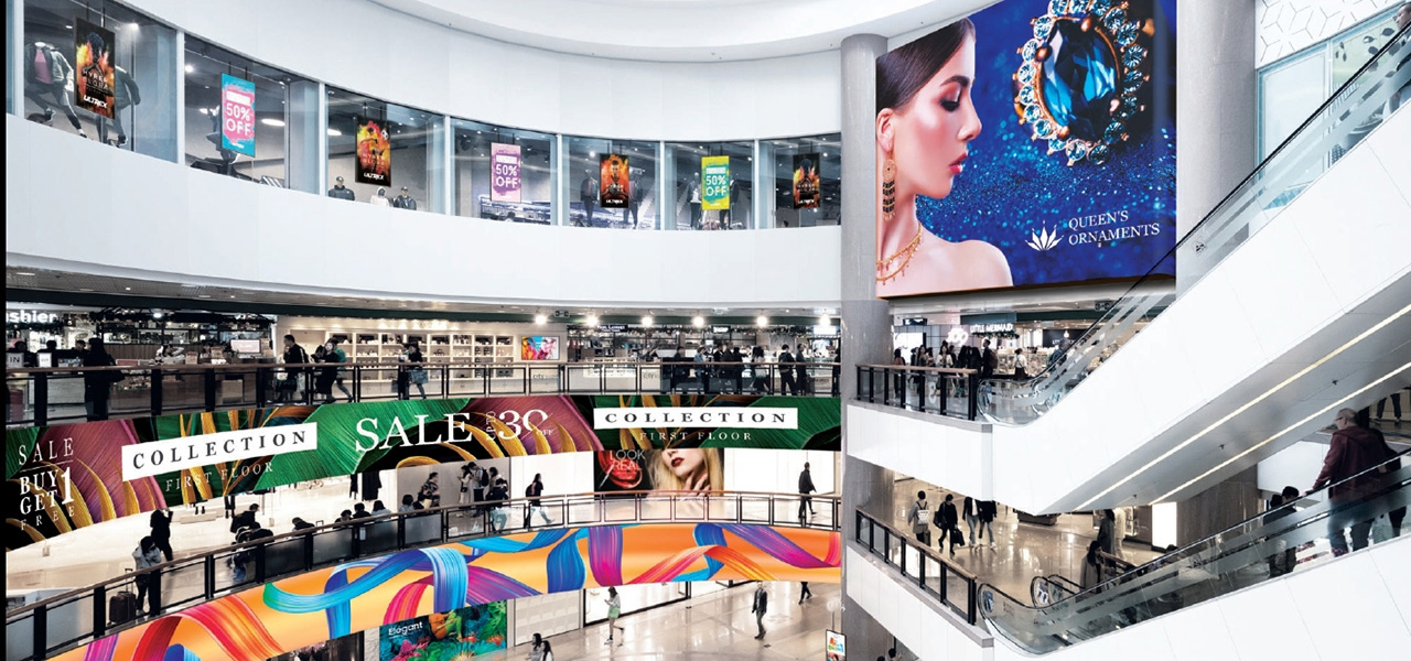 Wielopoziomowa galeria handlowa z ruchomymi schodami, licznymi ekranami reklamowymi nad witrynami sklepów i dużym wyświetlaczem z reklamą biżuterii