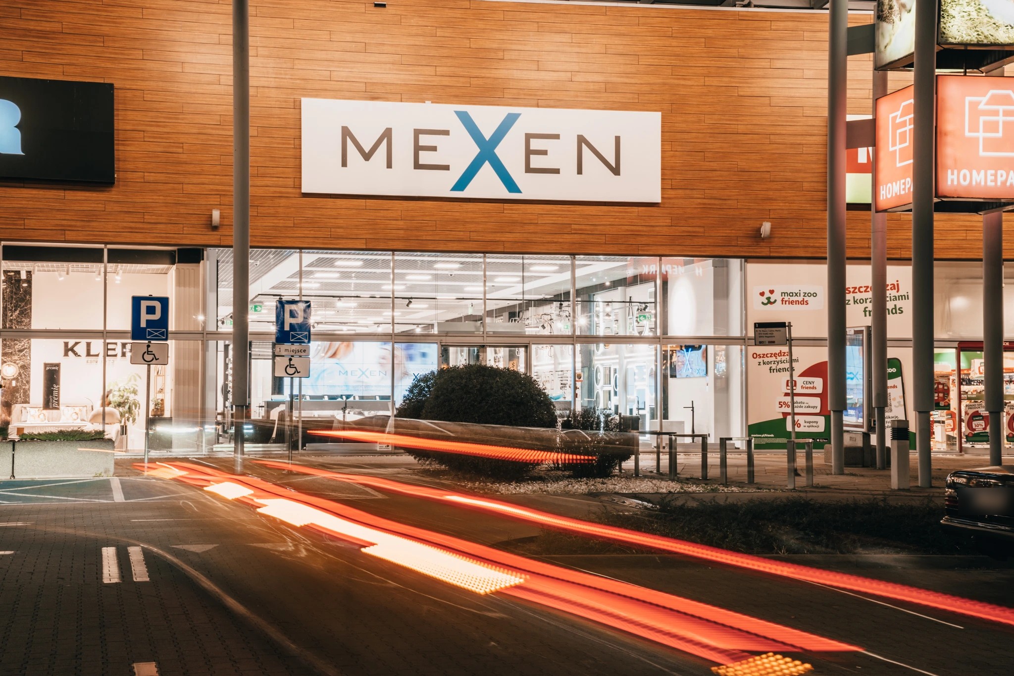 Fasada salonu Mexen w HOMEPARK Targówek wieczorem: podświetlone logo nad przeszklonym frontem, przed budynkiem smugi świateł przejeżdżających samochodów